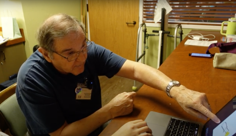 An older man wearing glasses and a navy-blue shirt sits at a wooden table, leaning forward and pointing at a laptop screen displaying a shiny fishing hook. His expression is focused and engaged as he gestures with his hand, demonstrating the ease and precision of an experienced angler.